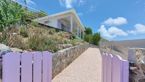 A pathway leading to a modern house with a white exterior and purple accents, surrounded by a variety of plants and a stone wall. - Villa Pinel – Intimate Oceanview Escape Above Orient Bay, St. Martin (Cul-de-Sac)