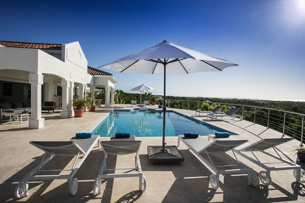Une piscine entourée de chaises longues et de parasols, le tout bordé d’un bâtiment blanc doté d’un toit en tuiles.