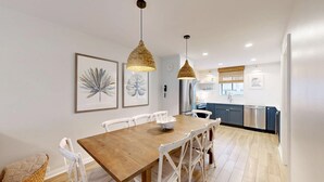A dining area with a wooden table and white chairs, a kitchen with blue cabinets, and two hanging pendant lights. - The Pool Is At Your Doorstep! (Hilton Head)