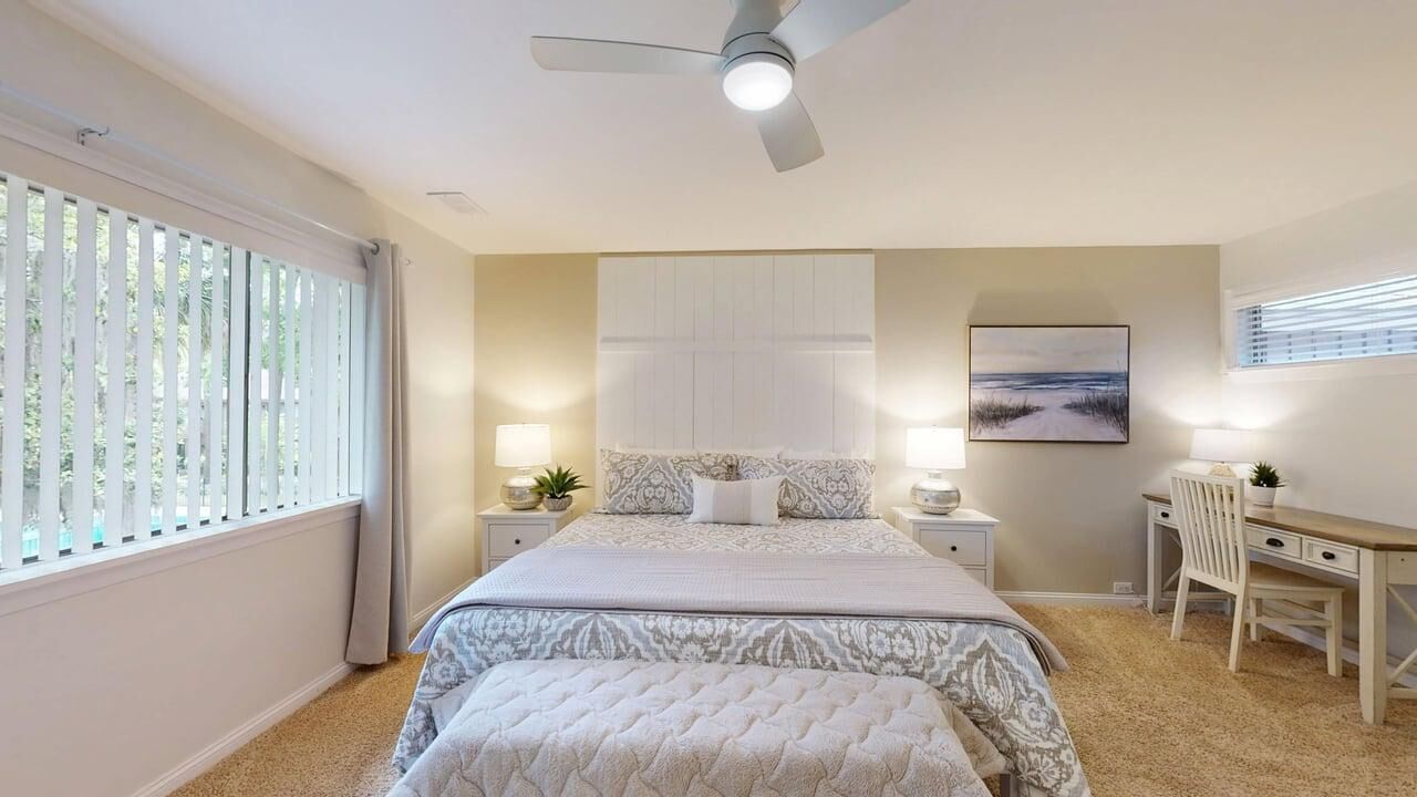A bedroom with a bed, bedside tables, a desk, a chair, a ceiling fan, and a window with blinds.