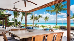 A wooden dining table with chairs, a bowl of fruit, and a view of the beach and ocean. - Hawksbill Estate – A Grand Beachfront Estate on Grace Bay, Turks & Caicos (Leeward Settlement)