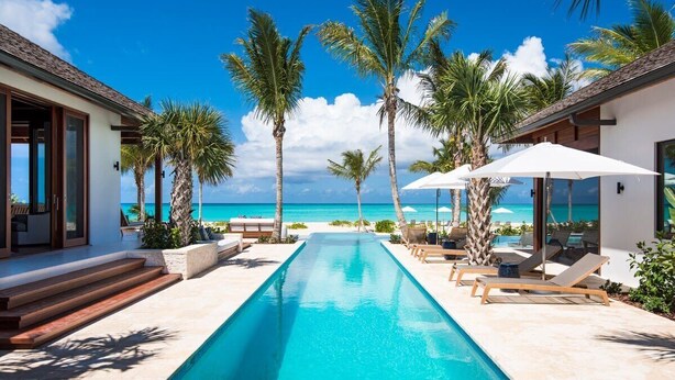 A swimming pool with lounge chairs and palm trees, leading to a beach. - Hawksbill Estate – A Grand Beachfront Estate on Grace Bay, Turks & Caicos (Leeward Settlement)