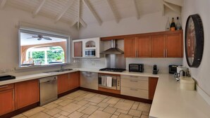 A modern kitchen with wooden cabinets, a built-in microwave, a stove, and a large window overlooking a green landscape. - Villa Sea Vous Play – Relaxed Oceanview Living in St. Martin (Cul-de-Sac)
