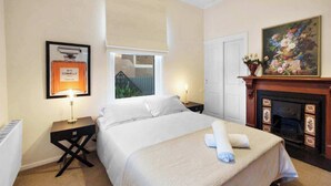 A neatly arranged bedroom with a bed, a nightstand, a framed perfume bottle, a window with a view, a fireplace, and a painting on the wall. - 5 Connelly - Echuca Holiday Homes (Echuca)