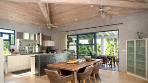 Een moderne keuken met een groot eiland, een eethoek met rieten stoelen, en een uitzicht op de omgeving door grote ramen.