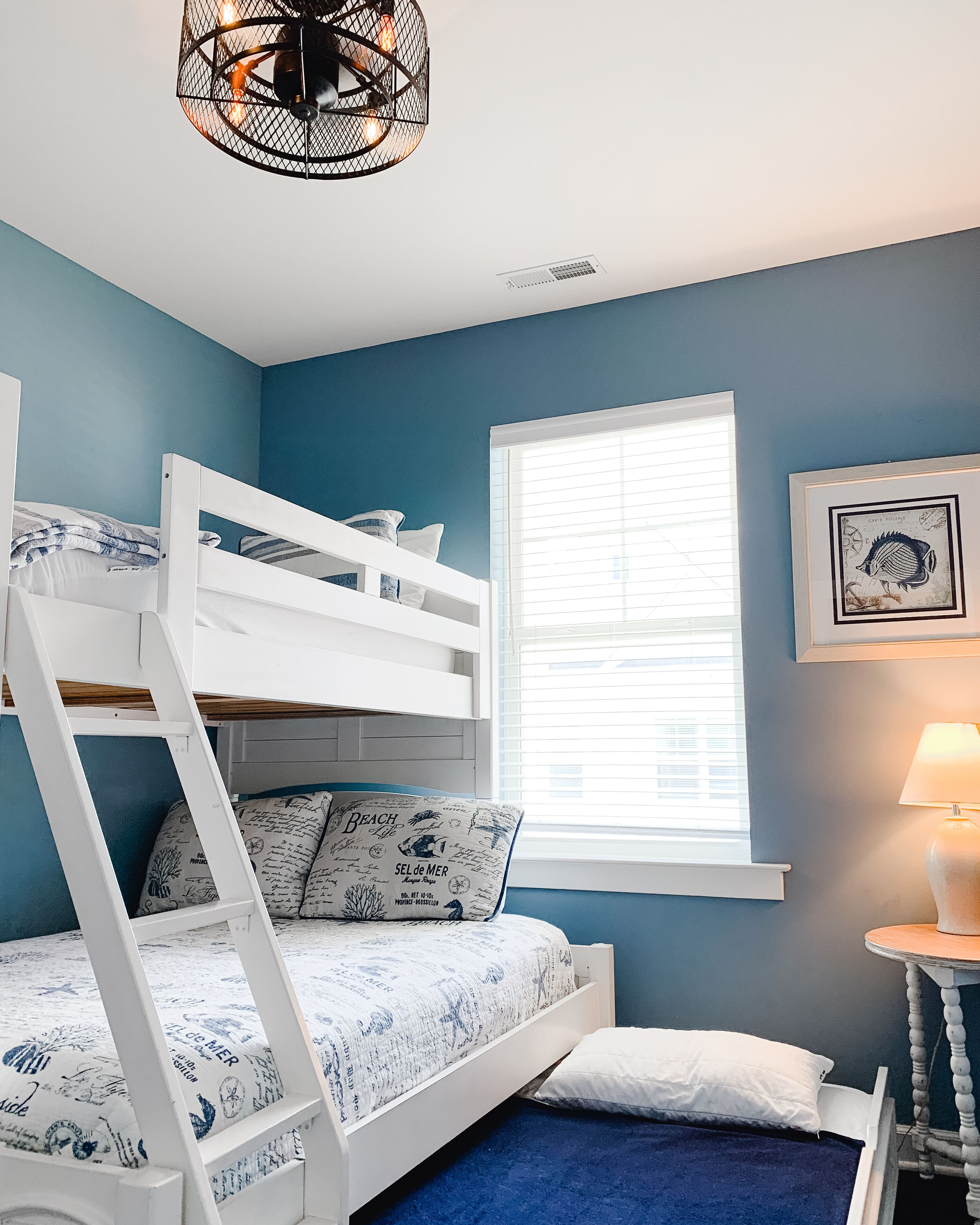 A bunk bed with a ladder, a window with blinds, a wall-mounted lamp, and a framed picture.