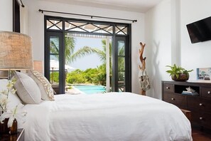 Ein Schlafzimmer mit Bett, Nachttisch, Lampe, Kommode und Blick auf einen Pool und Palmen.
