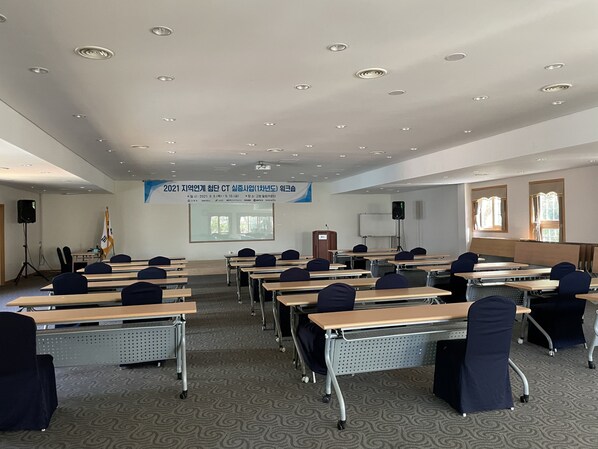 Un salón de conferencias con filas de escritorios y sillas, una pantalla de proyector y un cartel en la pared.