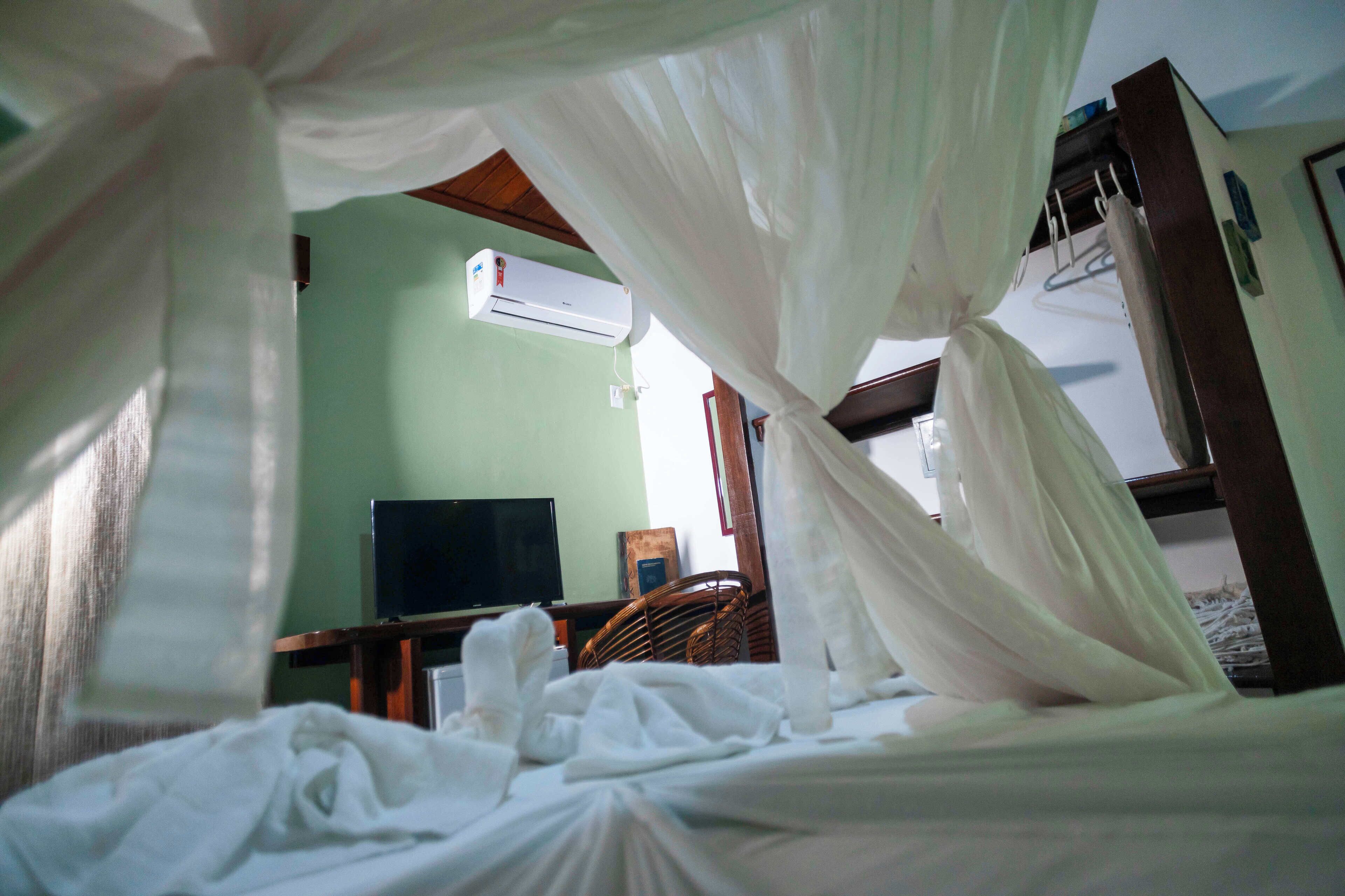 Quarto com cama coberta por um dossel branco, ar condicionado na parede, televisão sobre um suporte e uma cadeira de madeira.