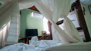Quarto com cama coberta por um dossel branco, ar condicionado na parede, televisão sobre um suporte e uma cadeira de madeira.
