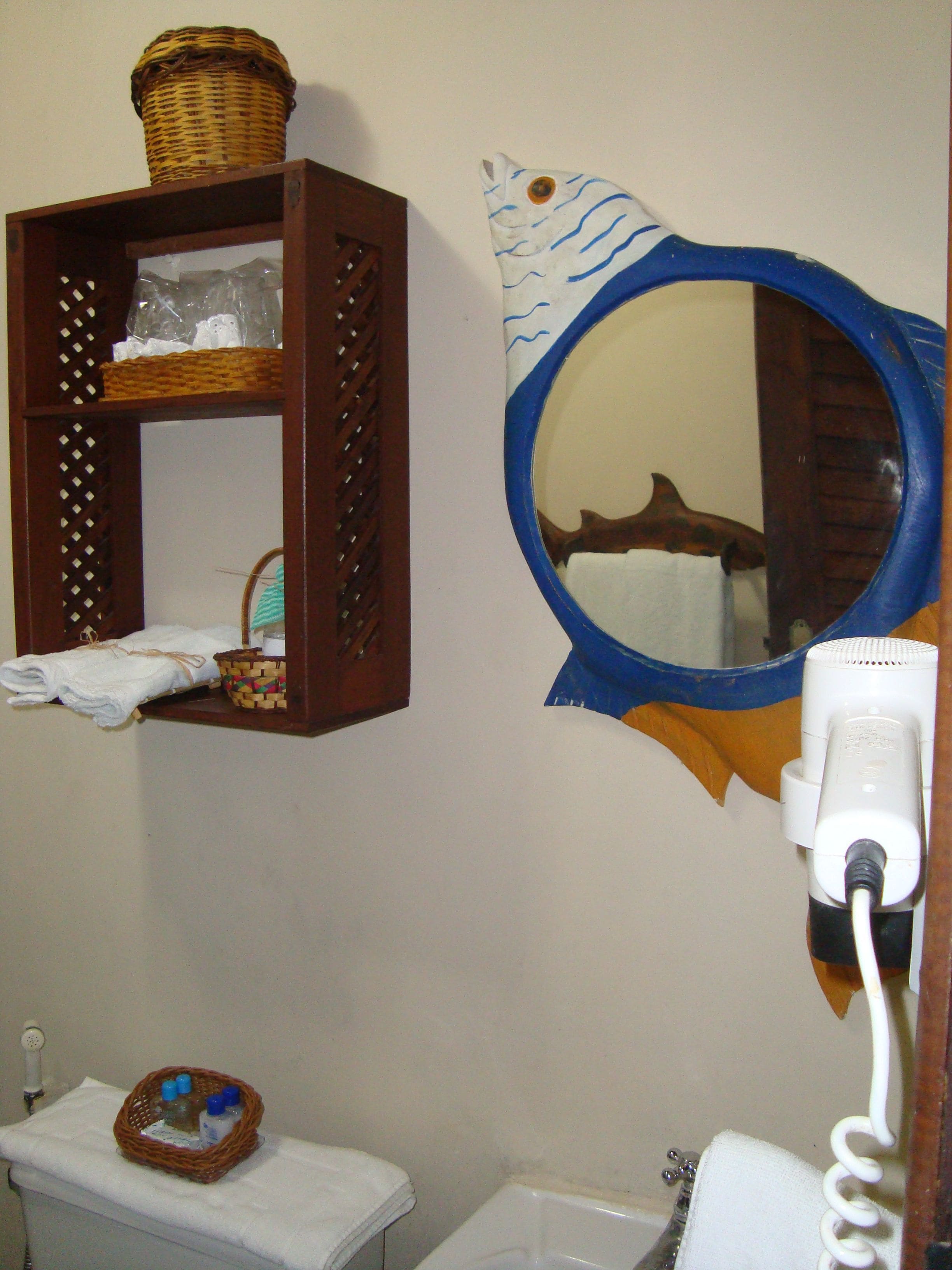 A bathroom with a wooden shelf holding a basket and a towel, a round mirror with a fish design, and a hairdryer plugged in.