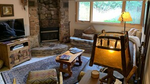 A cozy living room with a stone fireplace, a television, a sofa, and a wooden chair. - Historic 1925 Pine Cone Inn (Sandia Park)