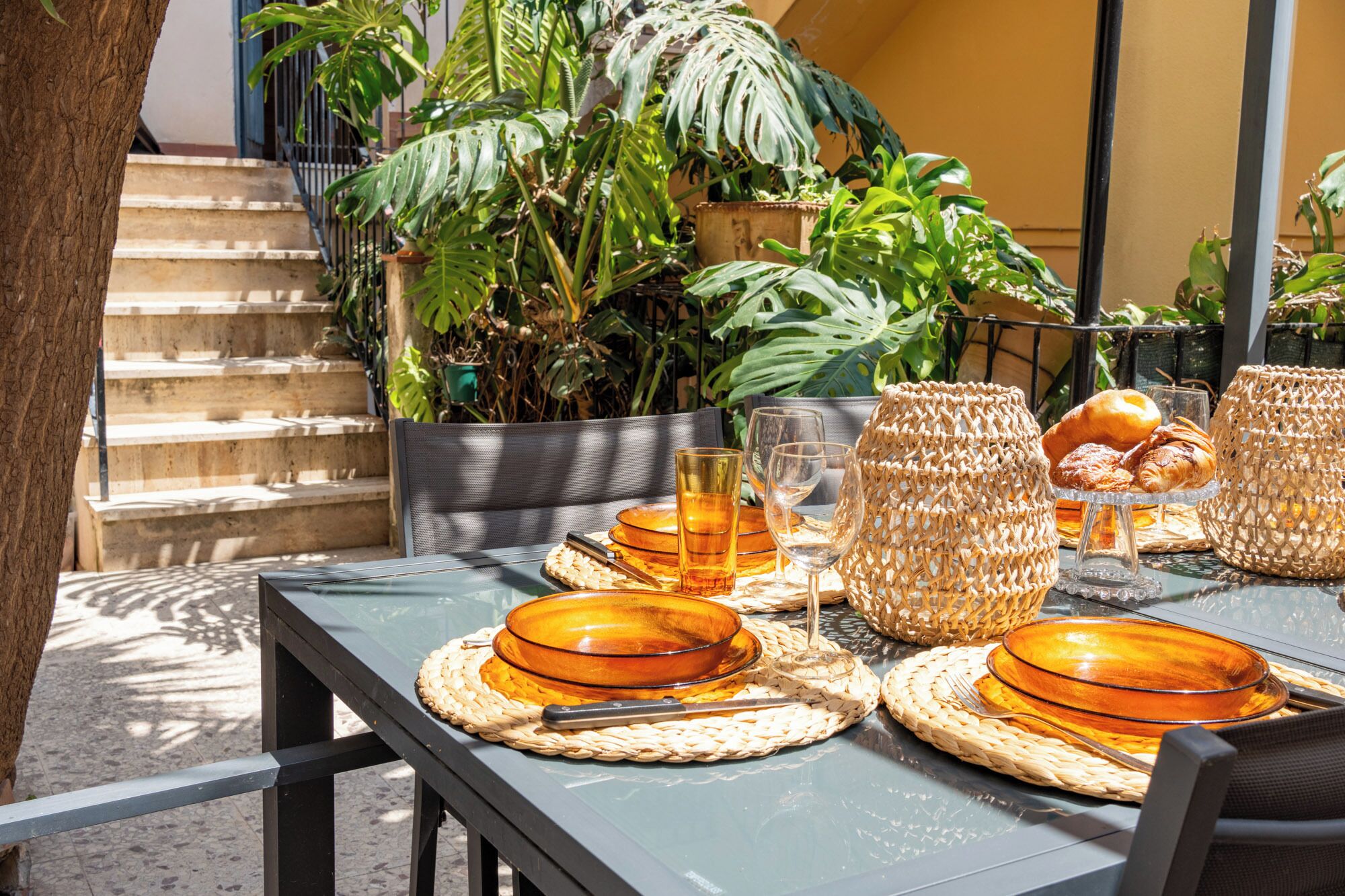 An outdoor dining area with a table set for two, surrounded by lush tropical plants.