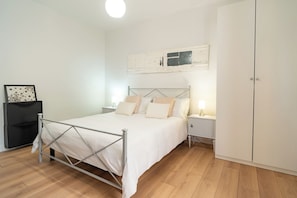 A bedroom with a metal-framed bed, white bedding, a bedside table with a lamp, a wardrobe, and a framed picture on the wall.