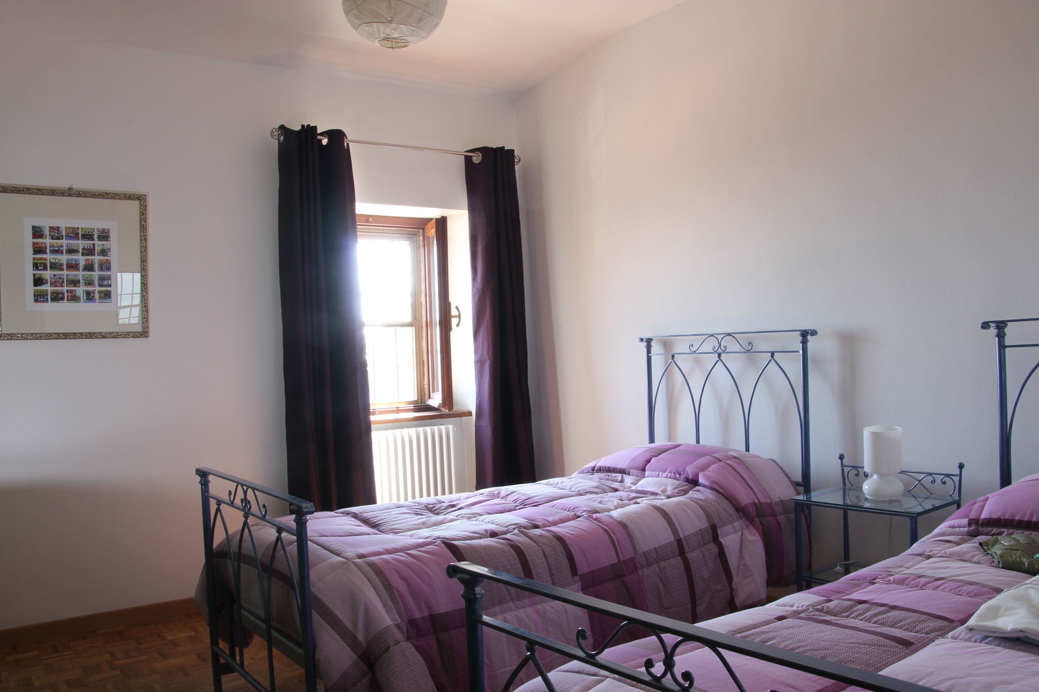 A bedroom with two beds, a window with curtains, a radiator, and a framed photo on the wall.