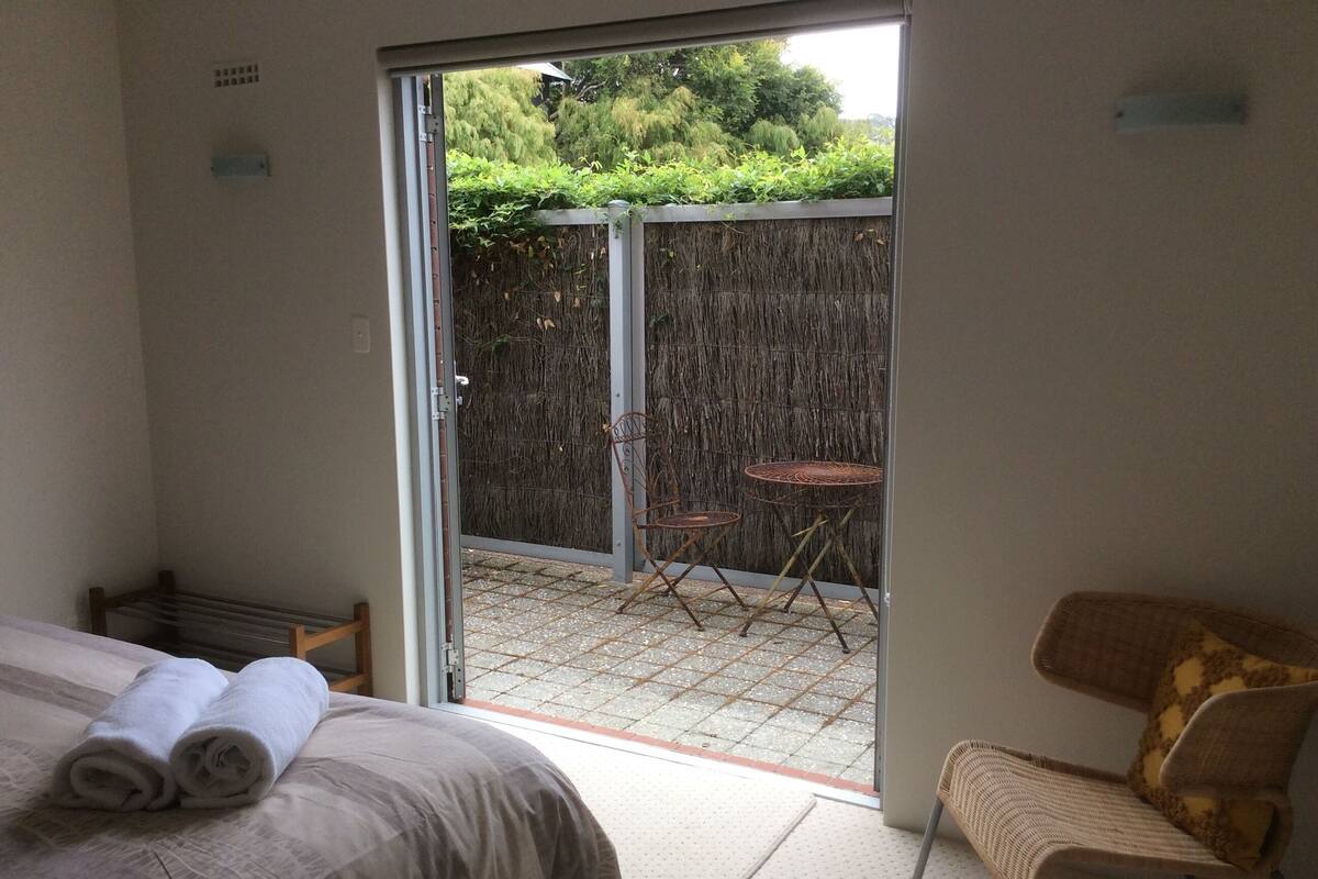 Ein Hotelzimmer mit einem Bett, einem Sessel, einem kleinen Tisch und einer Glastür, die zu einem Außenbereich mit Tisch und Stühlen führt.