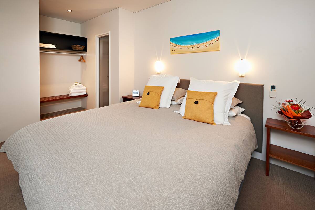 Ein ordentlich bezogenes Bett mit einer beigen Tagesdecke und zwei Kissen, ein Nachttisch mit einer Blumenvase, eine Wandlampe und ein Bild von einer Strandlandschaft.