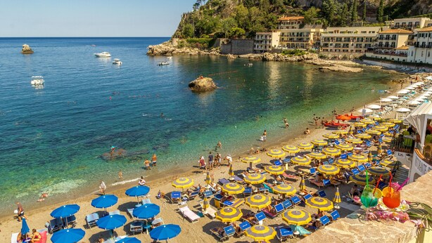 Una playa con agua cristalina azul, numerosas sombrillas y gente disfrutando del mar.