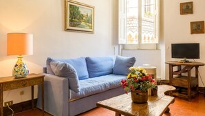 Una sala de estar con un sofá azul, una mesa de centro de madera con un jarrón de flores, una lámpara y un televisor sobre un mueble de madera.