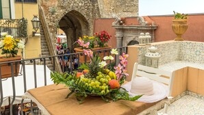 Un balcón con una mesa dispuesta con frutas y flores, una silla blanca y vistas a un edificio de piedra con un arco.