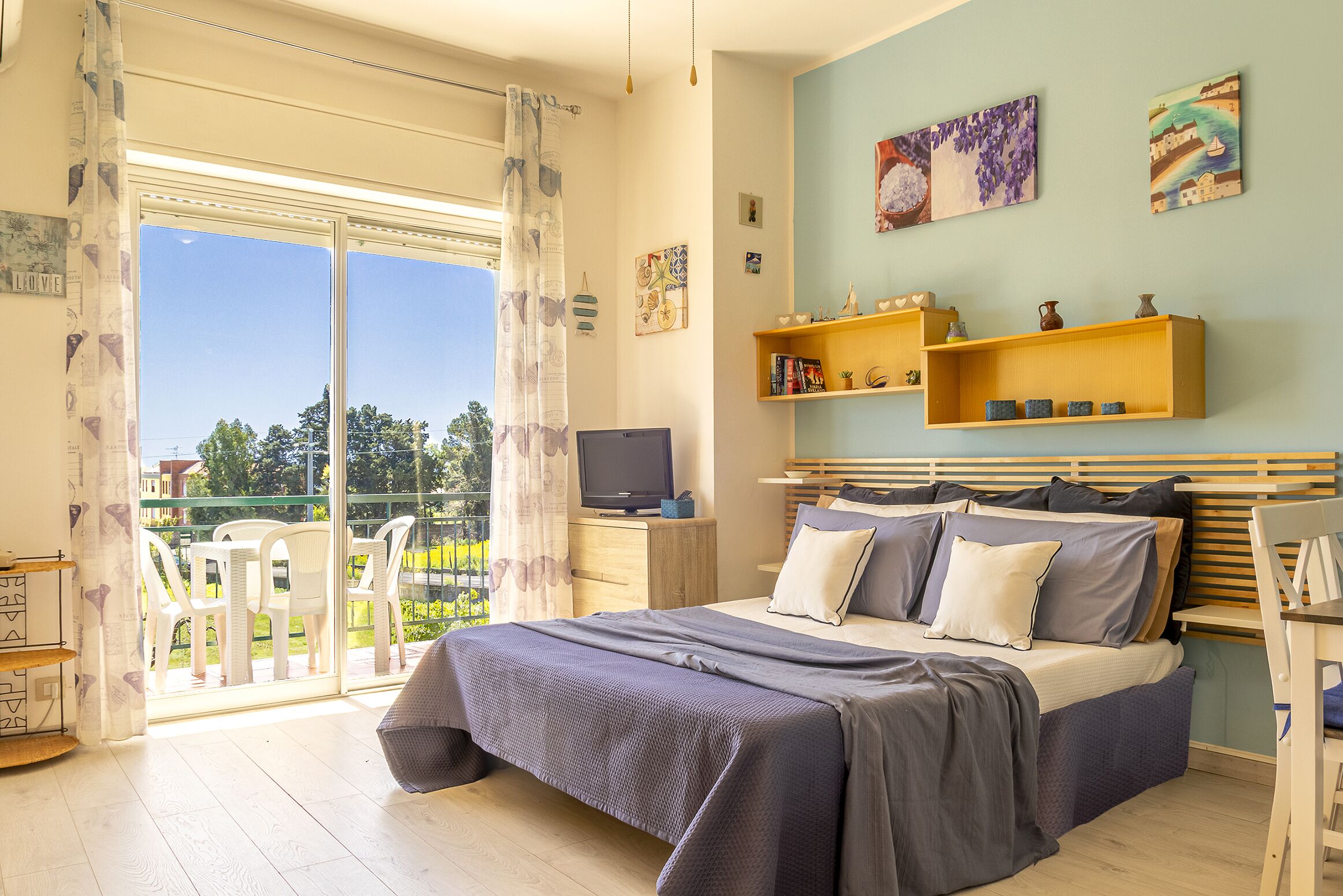 Un dormitorio con cama, televisor, balcón con sillas y obras de arte en las paredes.
