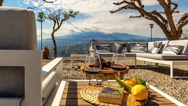 Un espace extérieur accueillant avec un canapé, une table agrémentée de verres à vin et d’un livre, et une vue sur les montagnes en arrière-plan.