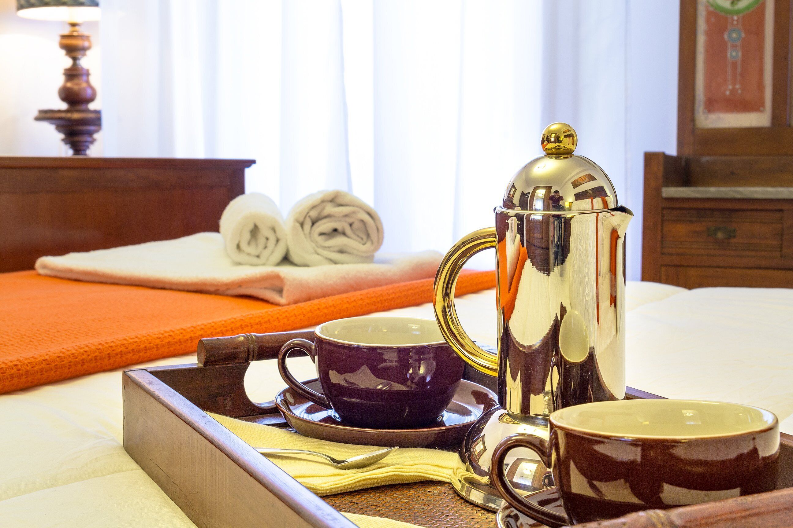 A neatly arranged breakfast tray with a coffee pot, two cups, and a spoon on a bed with a rolled towel and a wooden nightstand in the background.