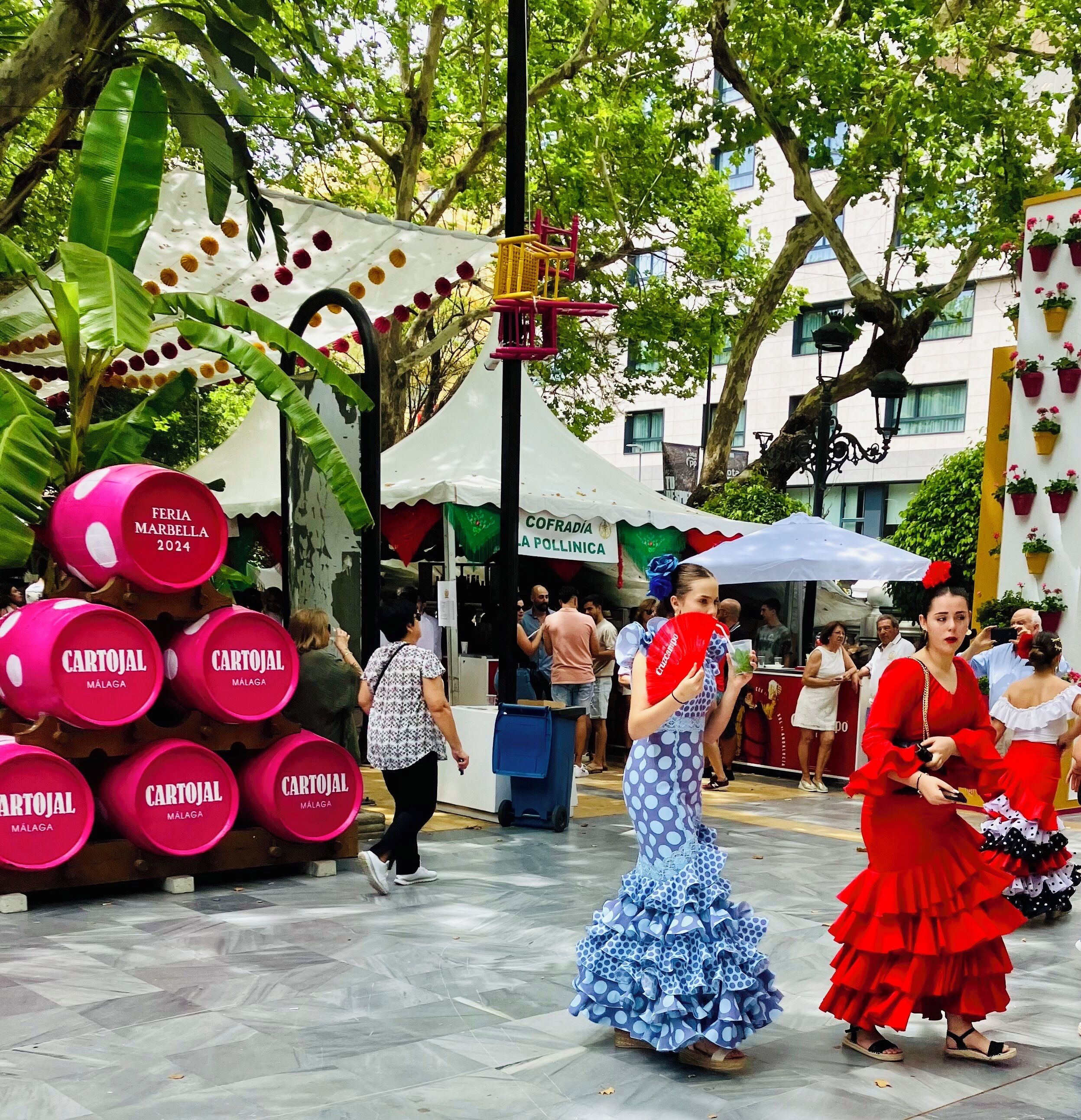 Ein öffentlicher Platz mit einem weißen Zelt, auf dem "COFRADIA A POLLINICA" steht, und einem Stand, der rosa Flaschen "CARTOJAL MALAGA" verkauft.