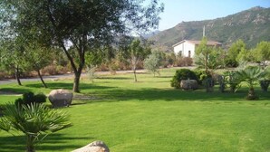Un área verde bien cuidada con rocas grandes y árboles, un edificio blanco al fondo y un paisaje montañoso.