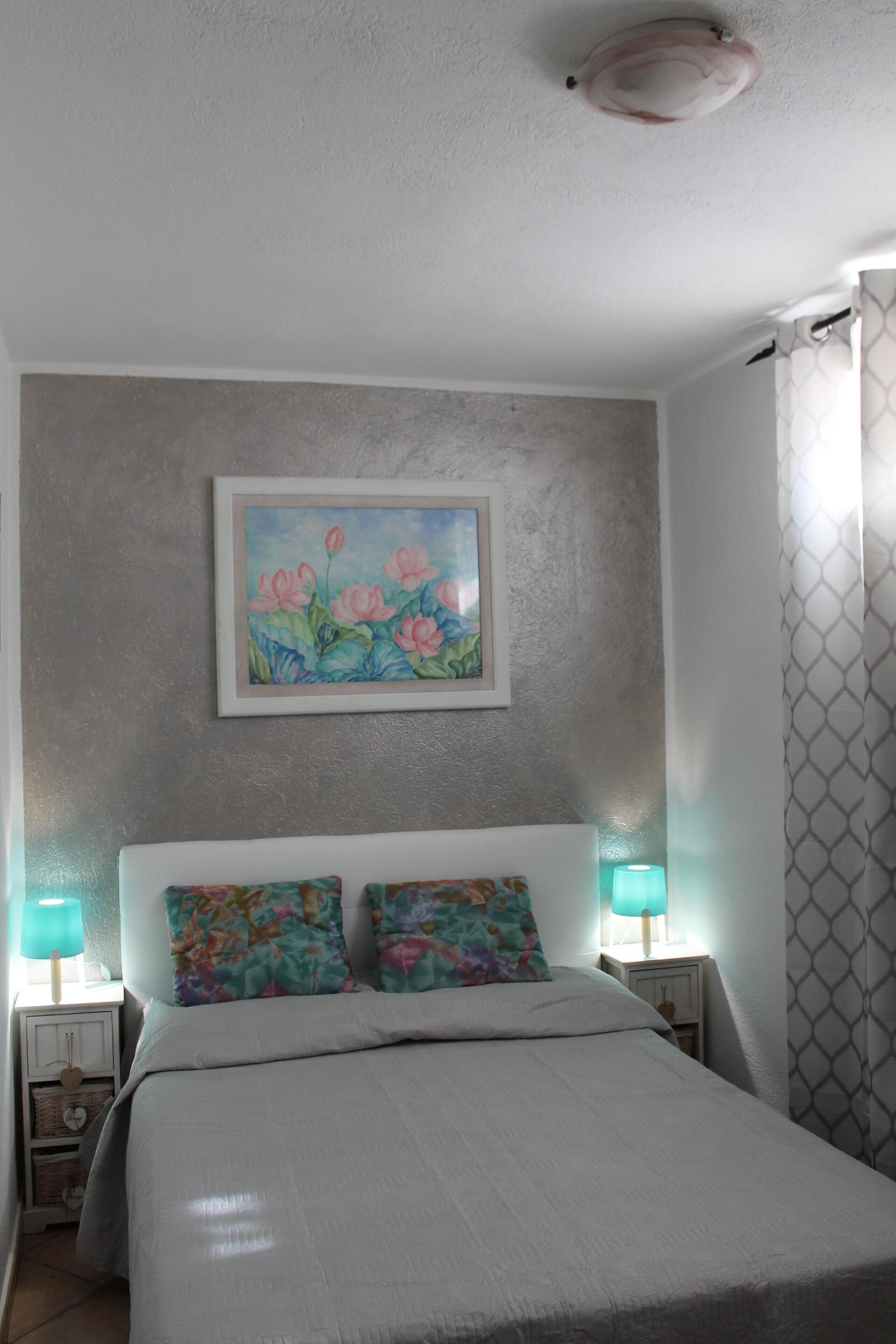 A neatly made bed with a white comforter and two colorful pillows, a wall-mounted painting of flowers, and two bedside lamps with teal shades.