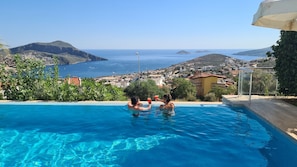 A swimming pool with clear blue water, surrounded by greenery and overlooking a coastal town with hills in the background.