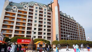 A multi-story hotel with balconies, a prominent tower, and a hotdog stand in the foreground. - Yangji Pine Resort (Yongin)