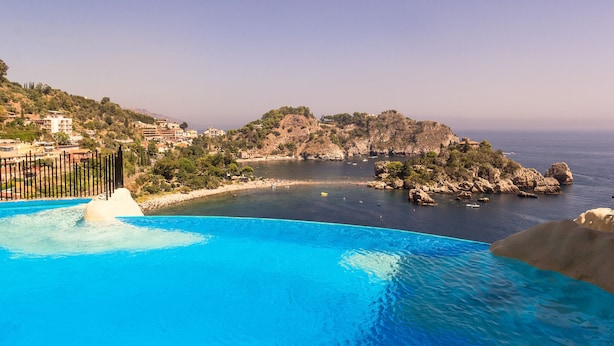 A coastal hotel with a swimming pool overlooking the sea, a rocky island, and a hillside with buildings. - Sea Life Taormina - Infinity pool (Taormina)