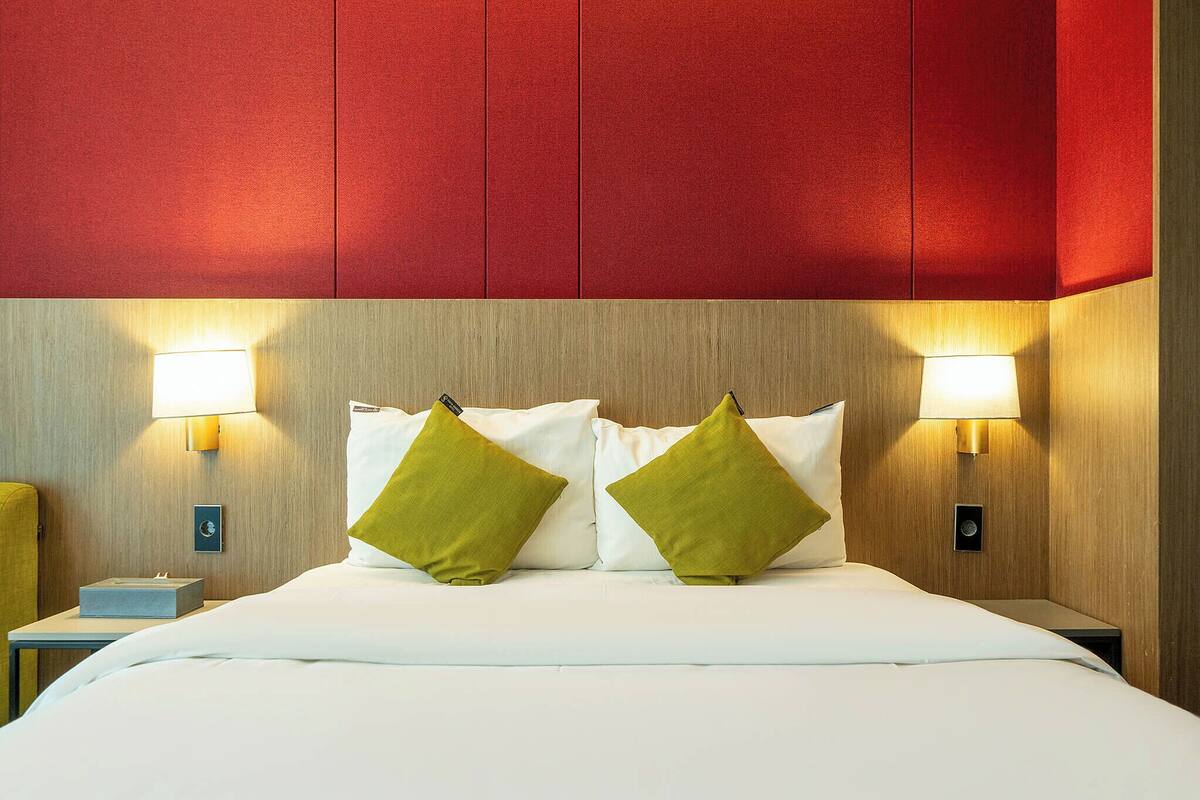 A hotel room with a bed, two green pillows, a bedside table with a lamp, and a nightstand with a lamp.