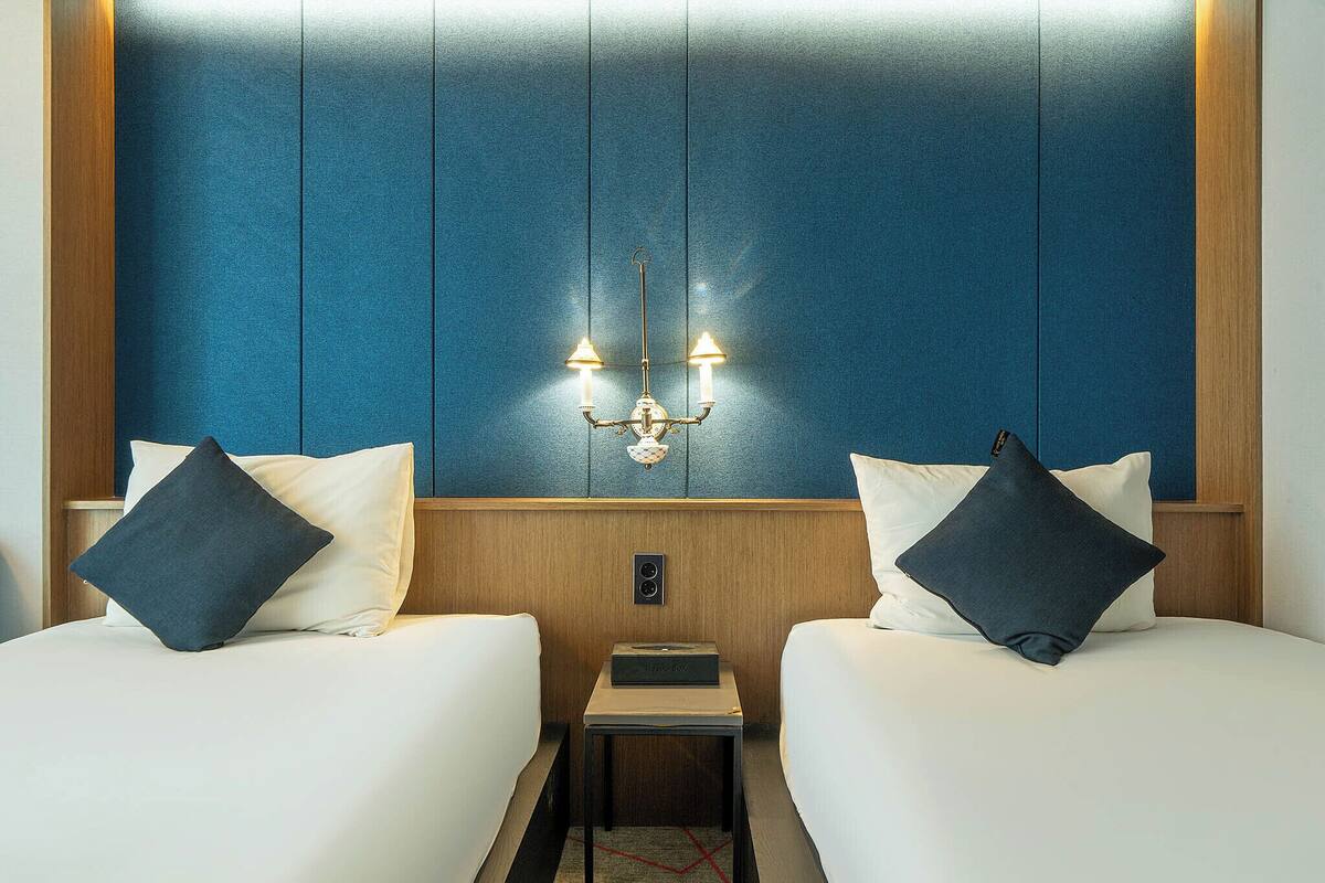 A hotel room with two beds, a nightstand, a wall-mounted light fixture, and a wooden headboard.