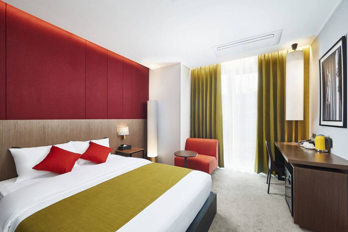 A hotel room with a bed, a desk, a chair, a lamp, and a large window with curtains.