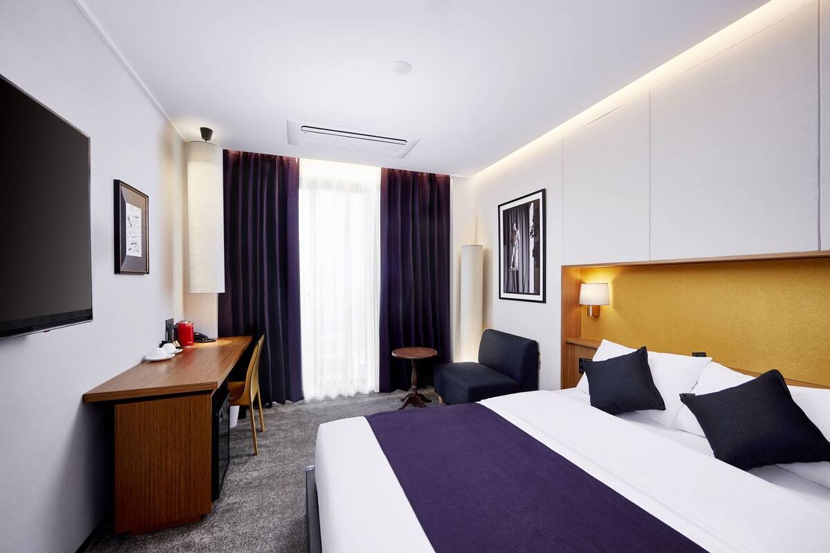 A hotel room with a bed, a desk, a chair, a small table, a TV, and a window with curtains.