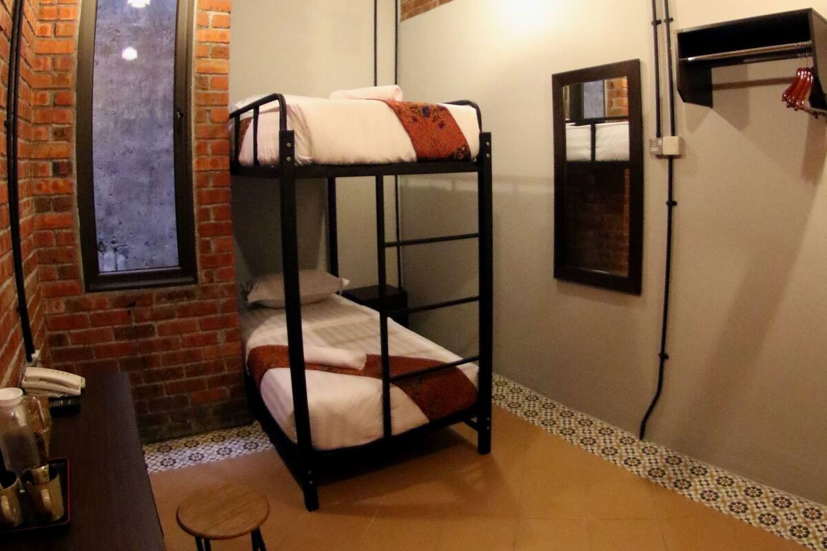 A bunk bed room with a brick wall, a small table with a chair, and a window.