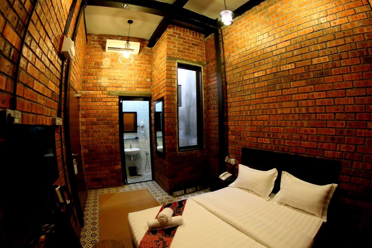 A hotel room with a brick wall, a bed with white linens, a small table, and a mirror.