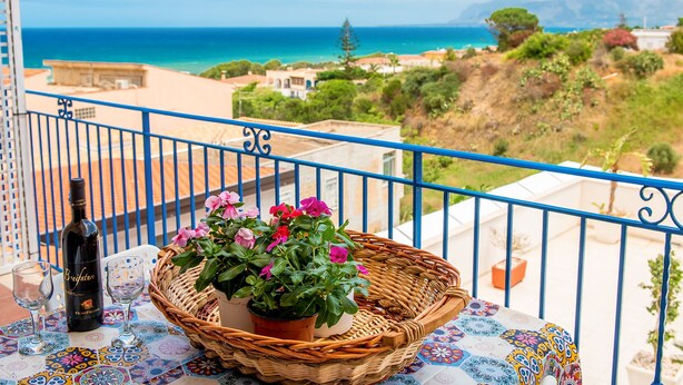 A balcony with a wicker basket containing pink flowers, a bottle of wine, and two glasses on a patterned tablecloth. - Apartment in villa wi fi 200 meters from the beach UA (Alcamo)