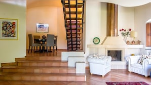 Una sala de estar con chimenea, sofás blancos, un comedor con mesa y sillas, y una escalera con barandilla de madera.