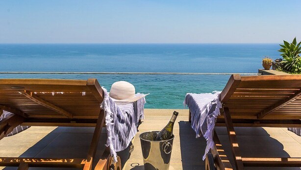 Una terraza frente al mar con sillones de madera, una botella de champán y una planta en maceta.