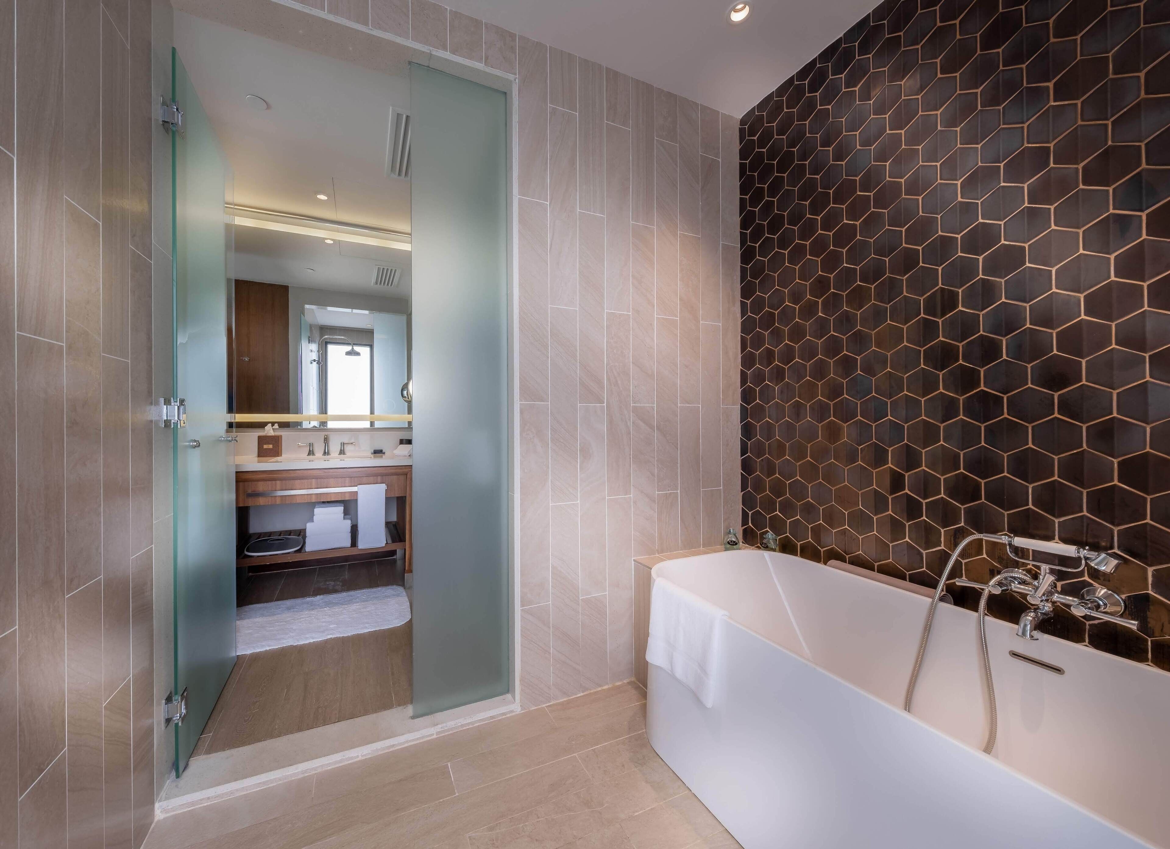 A modern bathroom with a glass door leading to a shower area, a large bathtub with a showerhead, and a hexagonal patterned wall.