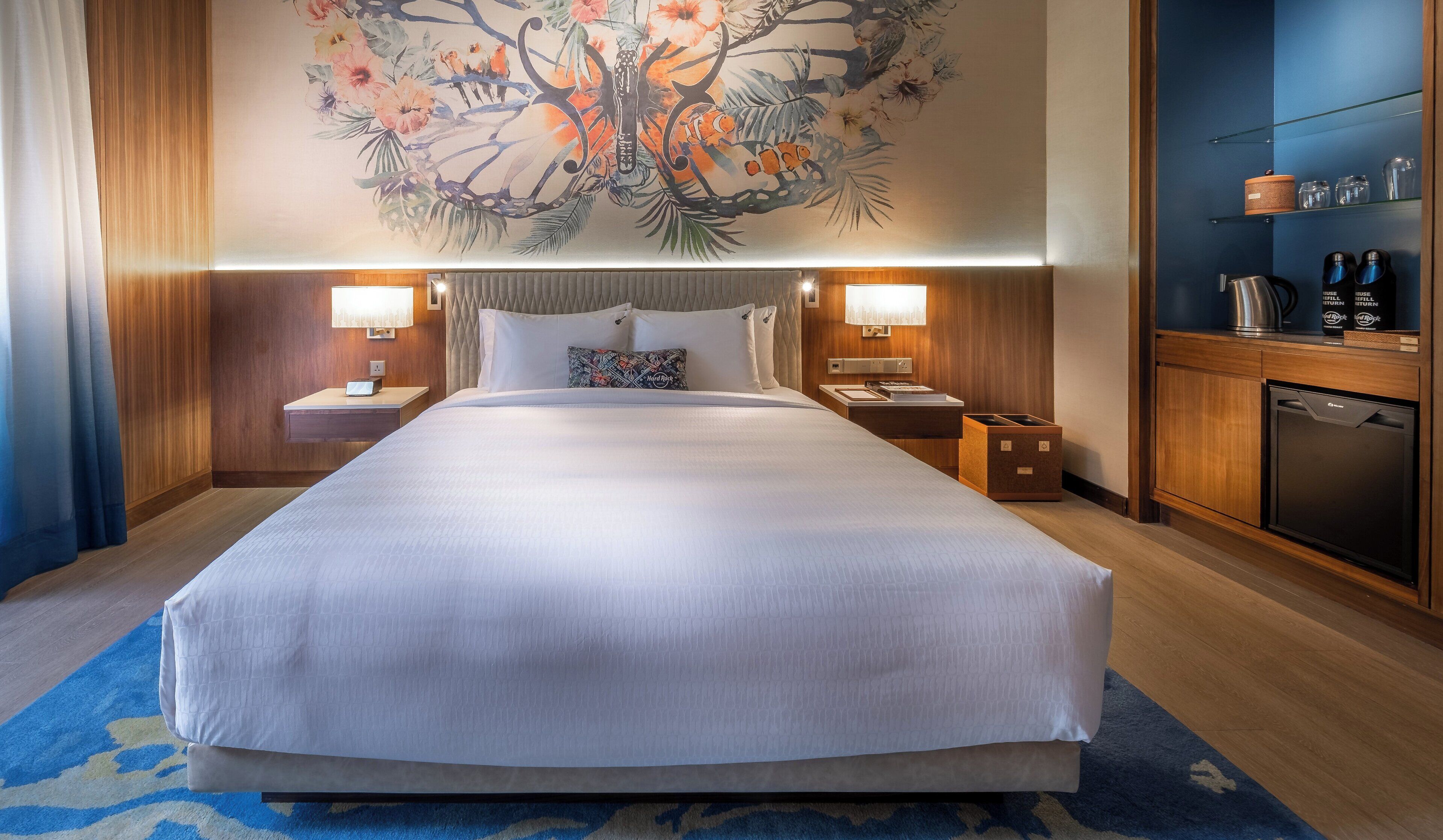 A large bed with white linens, two bedside tables with lamps, a nightstand, a wall-mounted shelf with glasses, a small refrigerator, and a decorative wall mural.