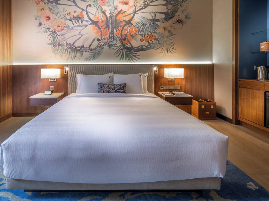A large bed with white linens, two bedside tables with lamps, a nightstand, a wall-mounted shelf with glasses, a small refrigerator, and a decorative wall mural.