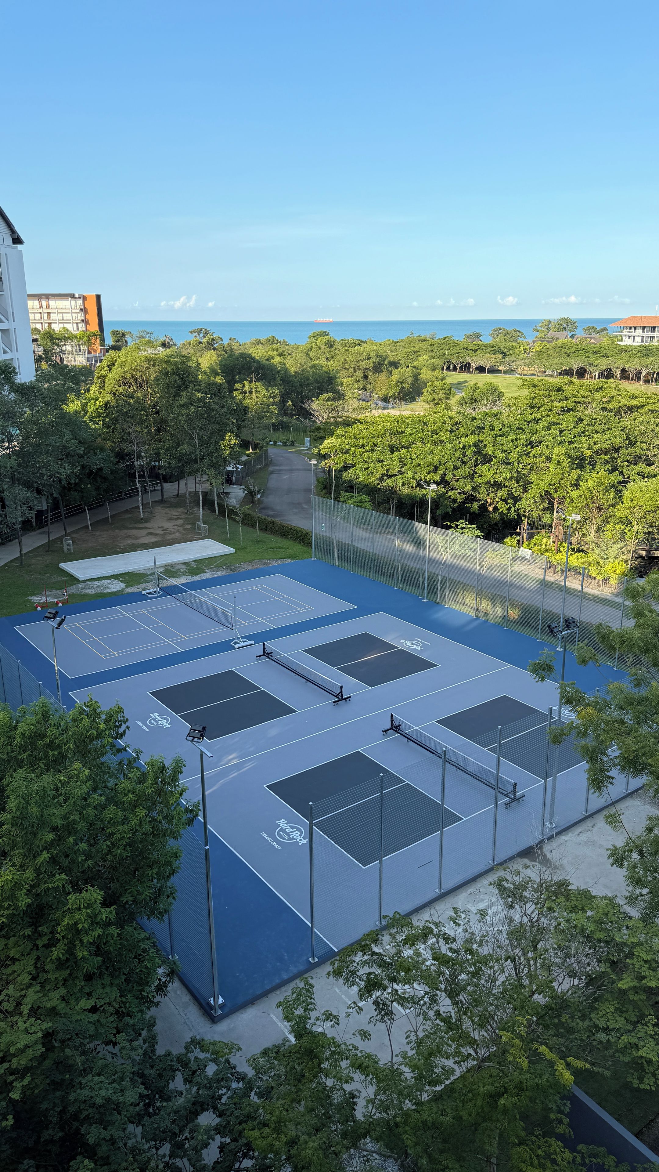 A tennis court with multiple courts, surrounded by trees and a view of the ocean.