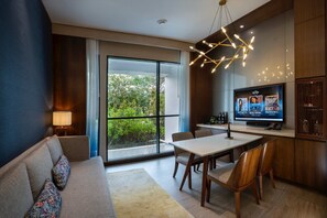 A modern living room with a sofa, a dining table with chairs, a TV, and a large window with a view of greenery.