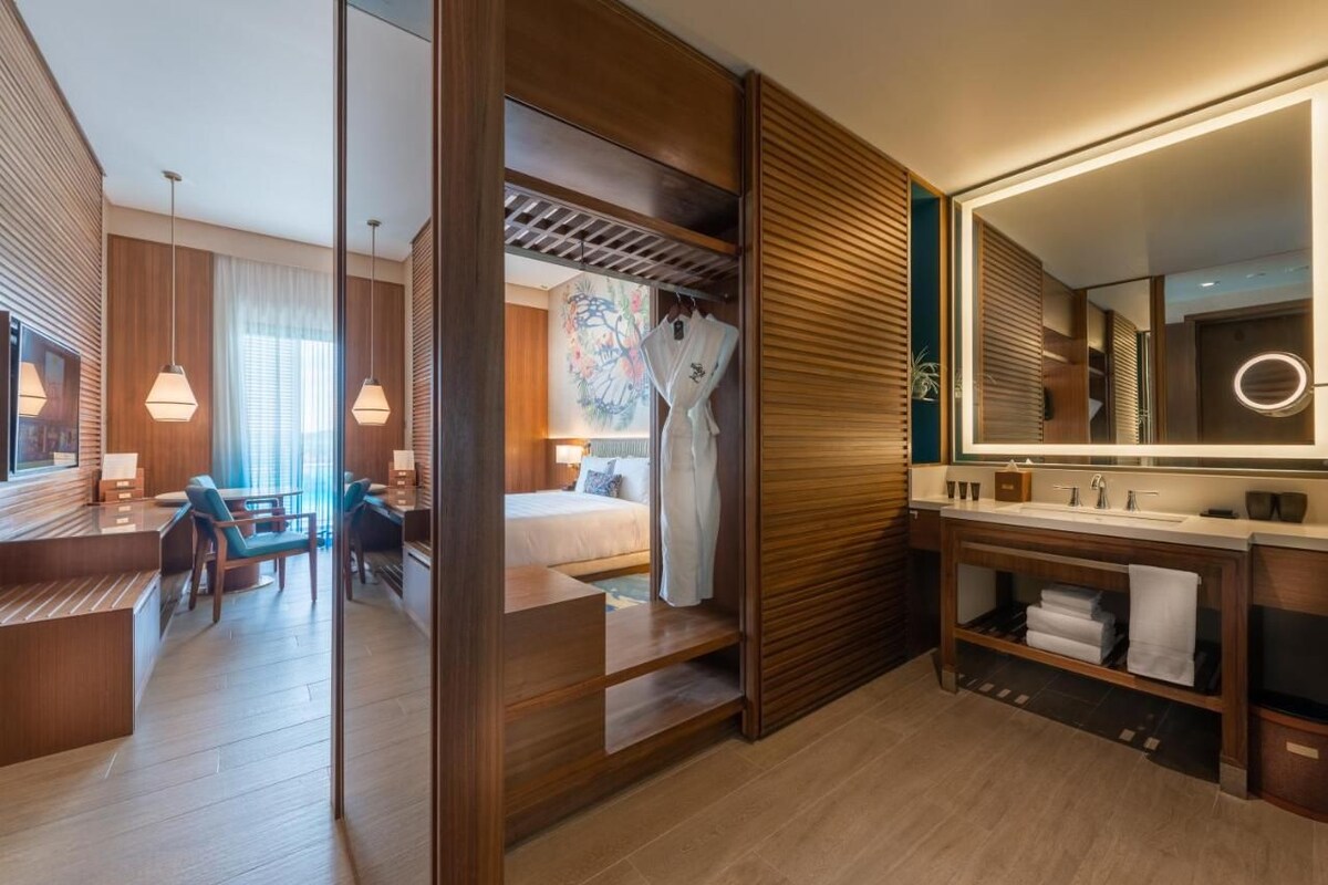 A modern hotel room with a bed, a desk, a chair, a bathroom with a mirror, and a window with curtains.