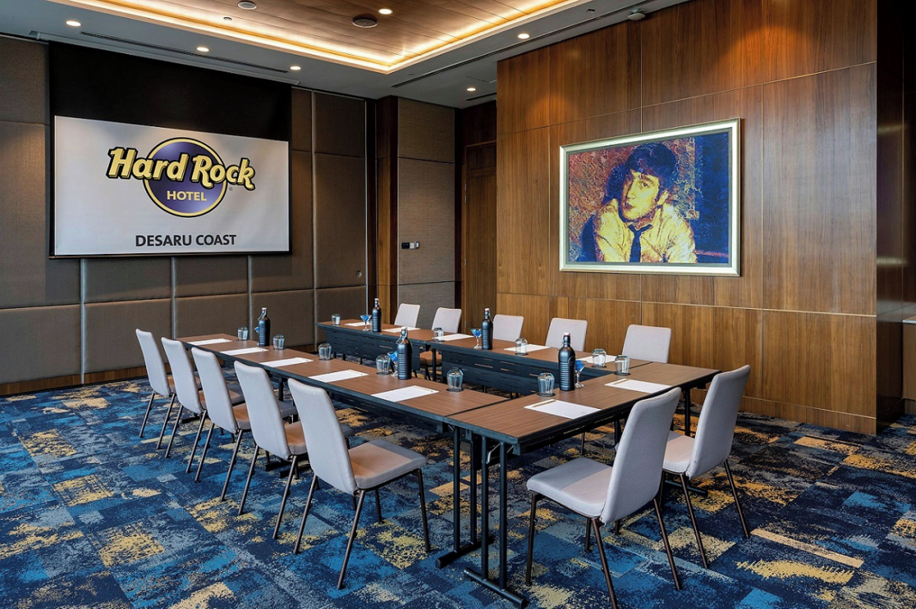 A conference room with a long table, chairs, and a projector screen displaying 'Hard Rock Hotel Desaru Coast'.