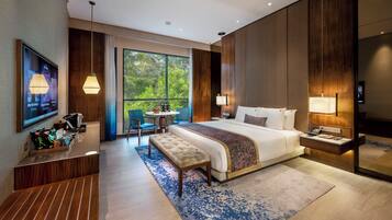 A modern hotel room with a large bed, bedside tables, a desk with a chair, a small round table with chairs, a bench, and a large window offering a view of greenery.